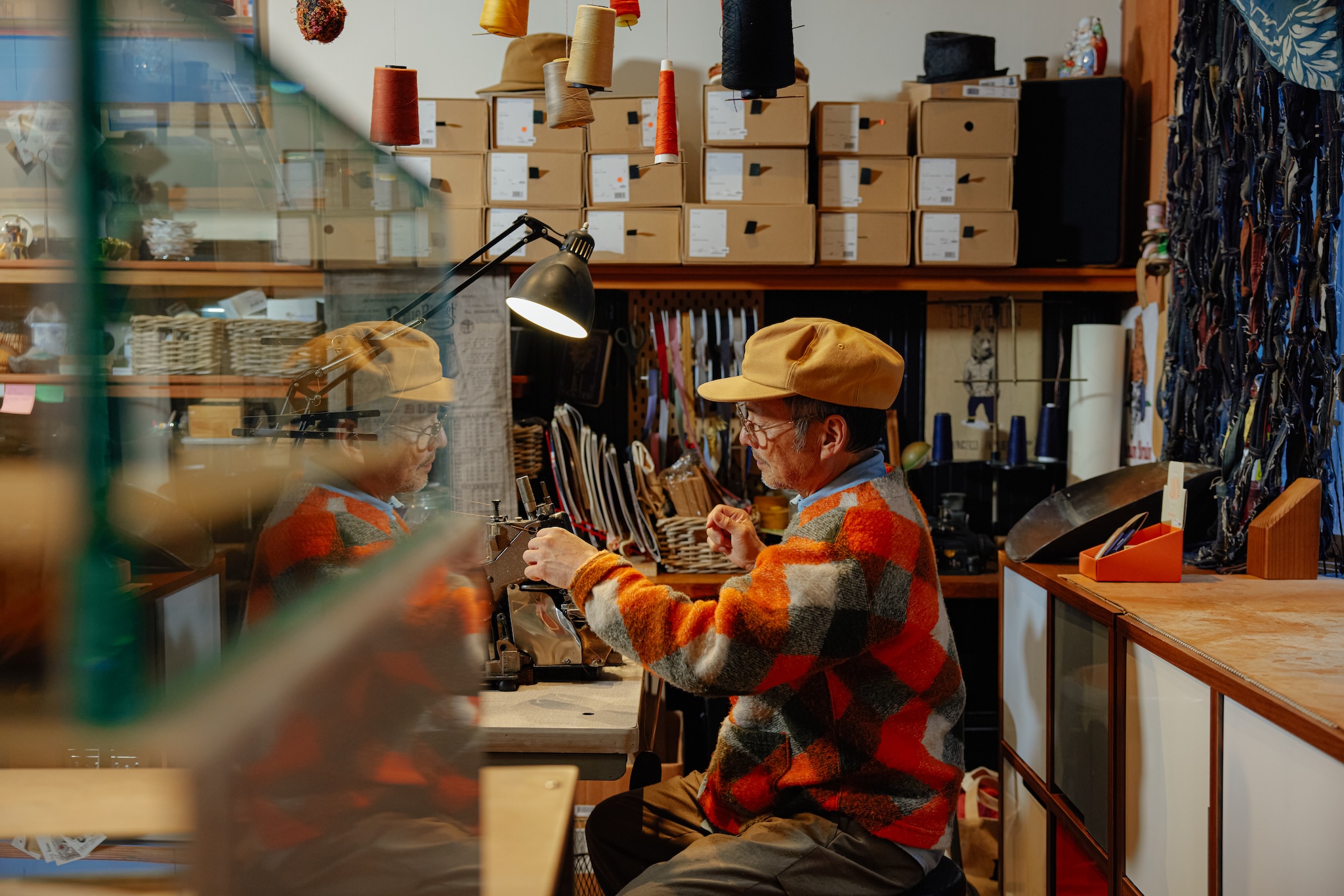 a stylish older man in a bright yellow cap and orange-checked sweater sits at his sewing machine
