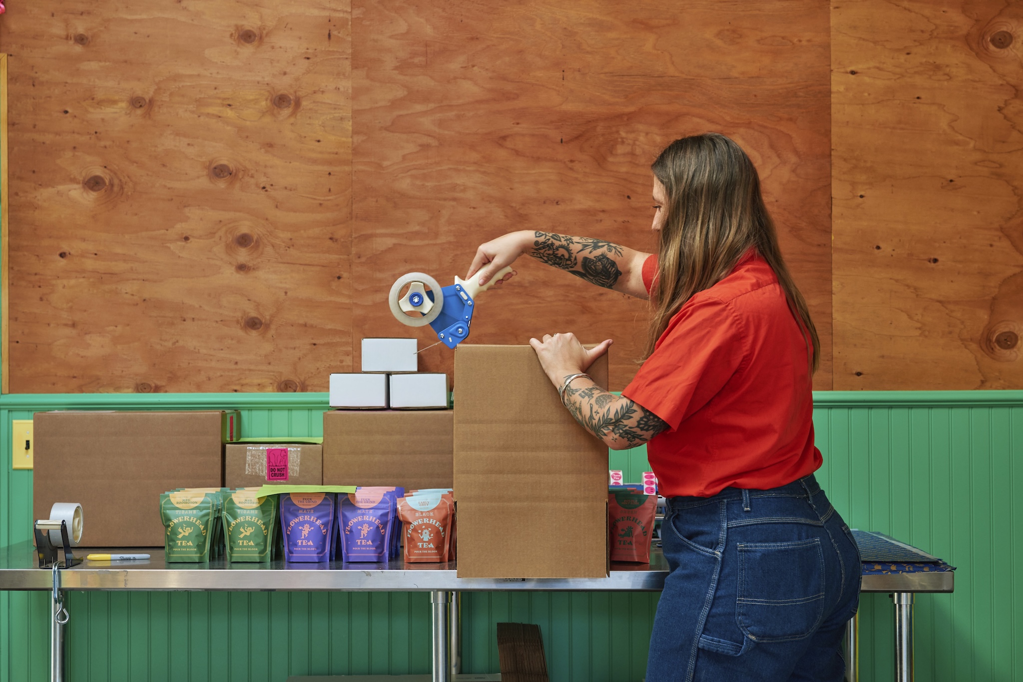A female worker in a bright red shirt is taping up cardboard boxes full of colorful packets of tea