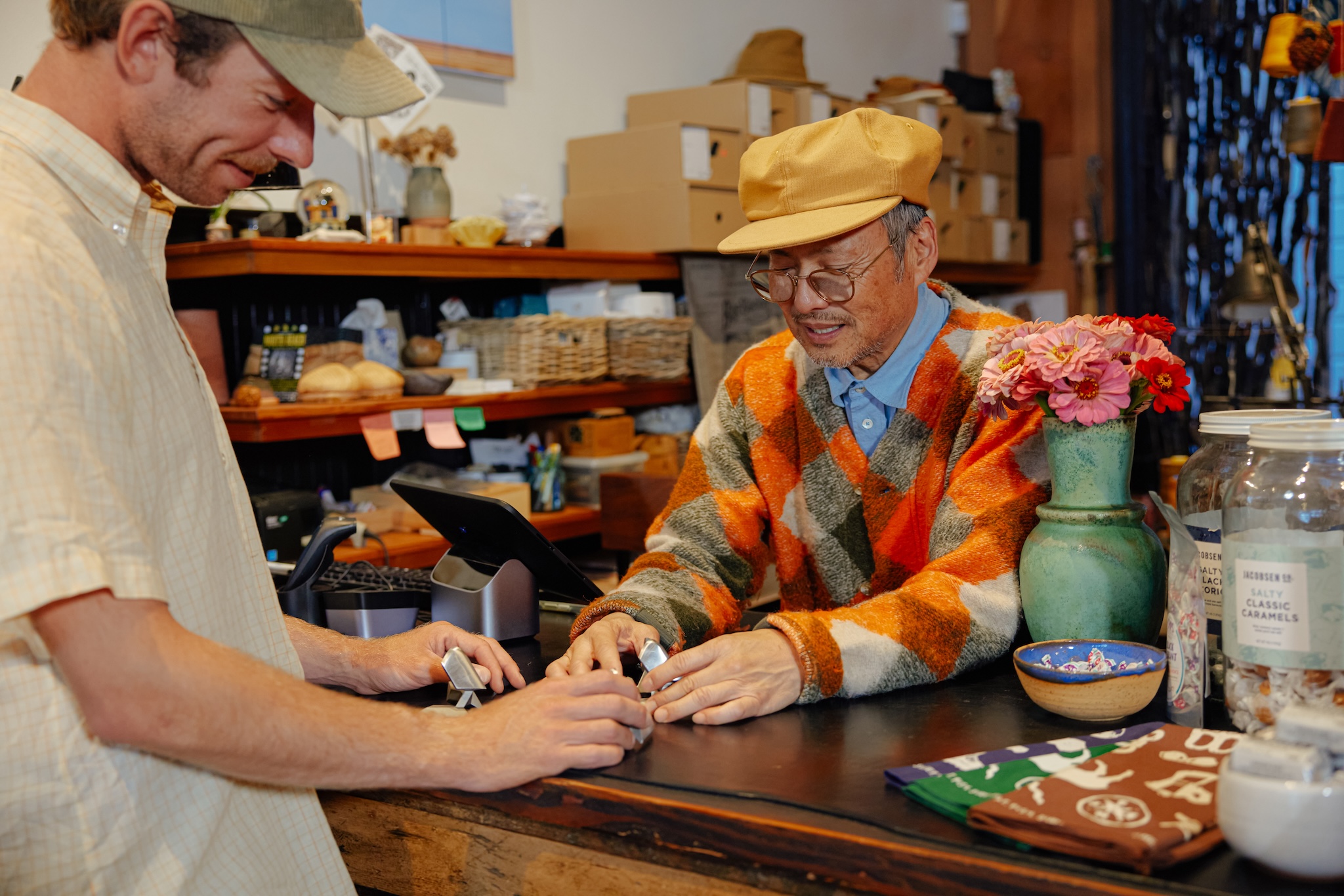 a stylish older male shopkeeper wearing a yellow hat checks out a customer at the cashier