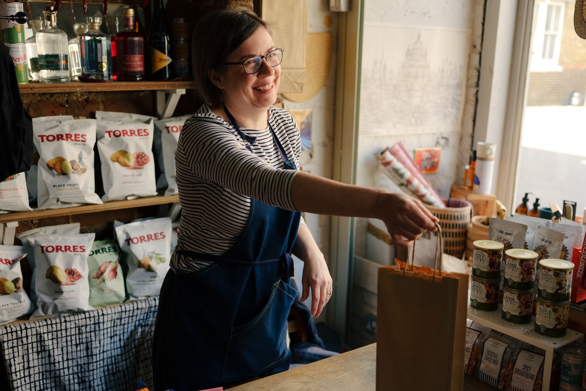 a smiling shopkeeper in a food retailer hands a brown paper bag across the counter